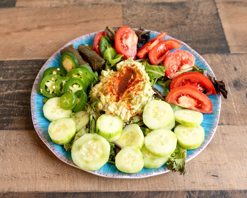 Colorful Mediterranean platter with hummus, tomatoes, cucumbers, jalapeños, and stuffed grape leaves.