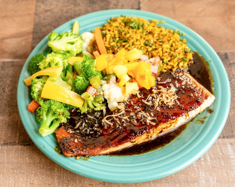 Grilled salmon with dark sauce, toasted coconut, yellow rice, and steamed vegetables on a teal plate.