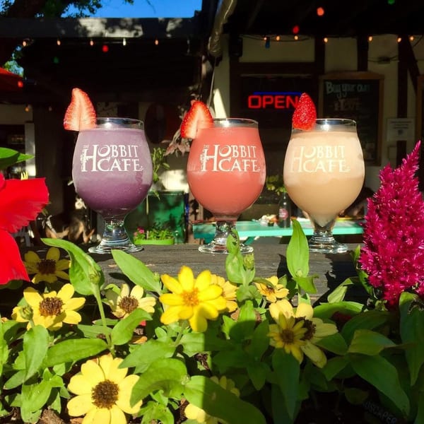 Three colorful Hobbit Cafe drinks with strawberry garnishes, framed by vibrant flowers on a sunny patio.