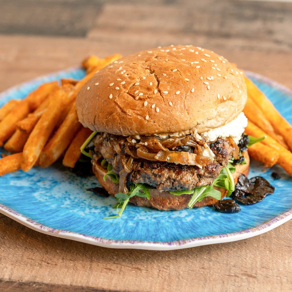 Gourmet burger with caramelized onions, goat cheese, and arugula, served with fries on a blue plate.