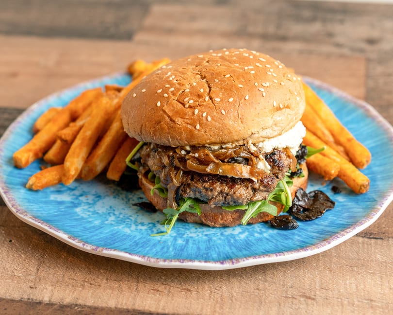 Gourmet burger with caramelized onions, goat cheese, and arugula, served with fries on a blue plate.