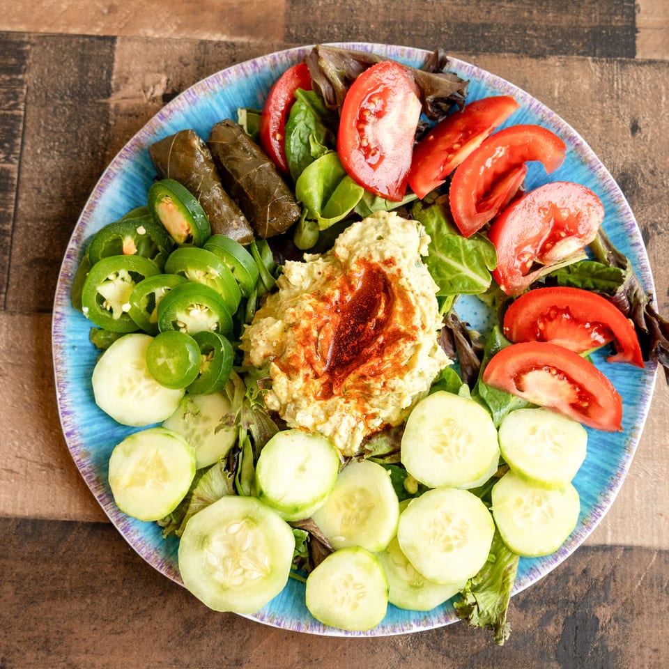 A colorful platter of hummus, stuffed grape leaves, sliced tomatoes, cucumbers, and jalapeños.