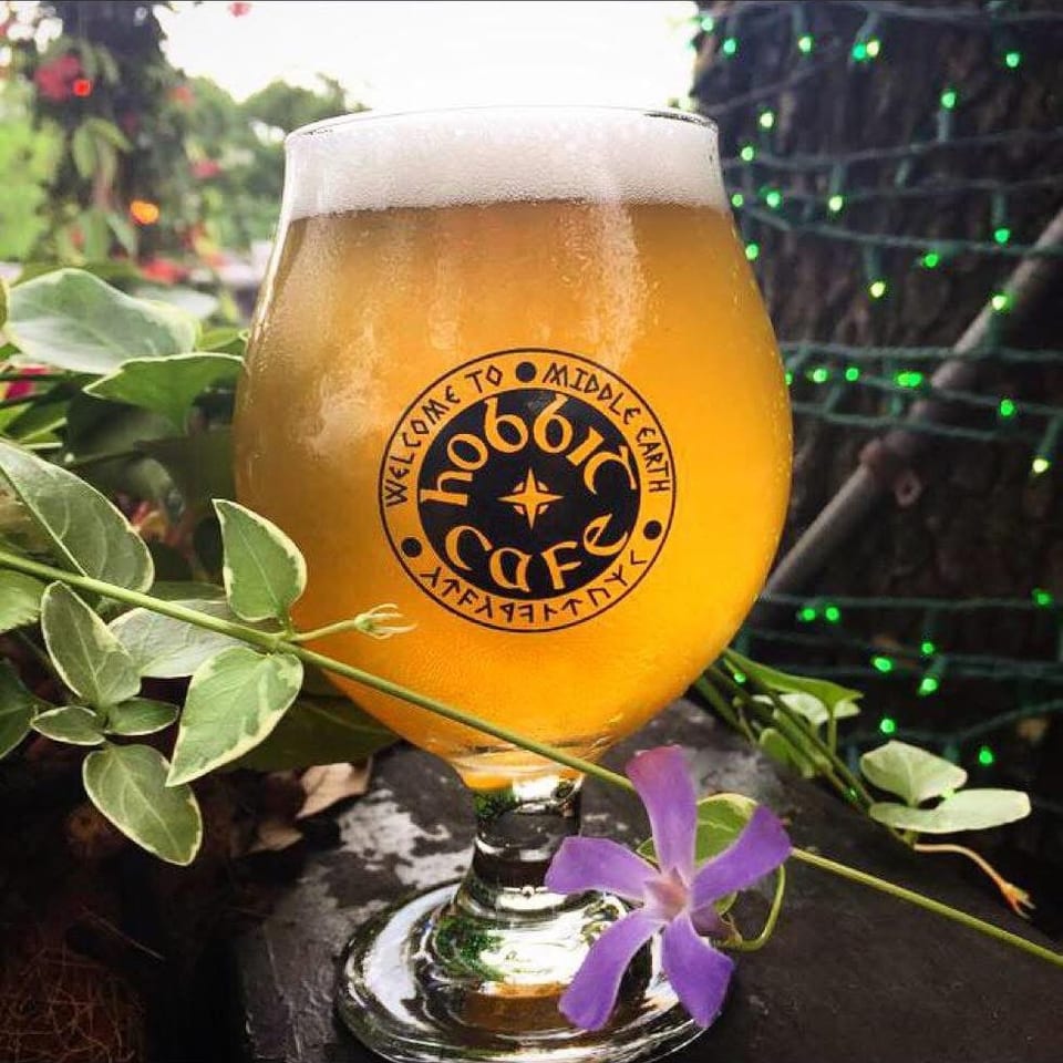 A glass of beer with "Hobbit Cafe" logo, surrounded by green leaves and a purple flower.