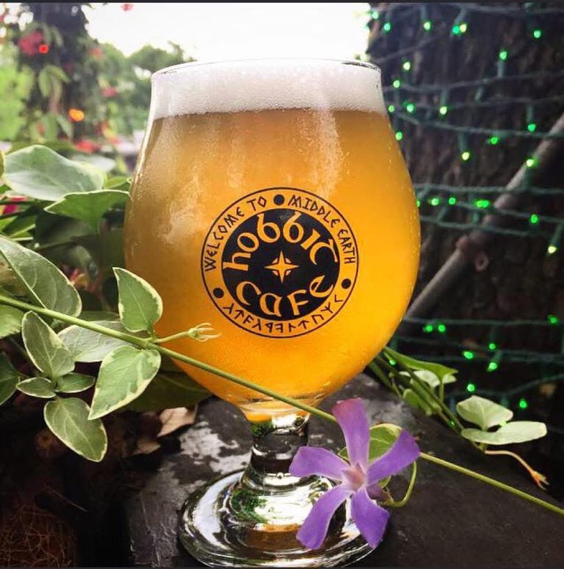 A glass of beer with "Hobbit Cafe" logo, surrounded by green leaves and a purple flower.