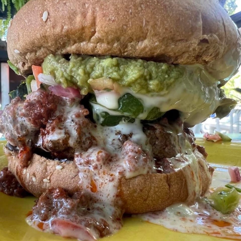 A large, messy burger overflowing with chili, melted cheese, and guacamole on a yellow plate.