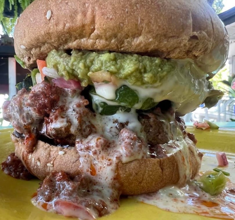 A large, messy burger overflowing with chili, melted cheese, and guacamole on a yellow plate.