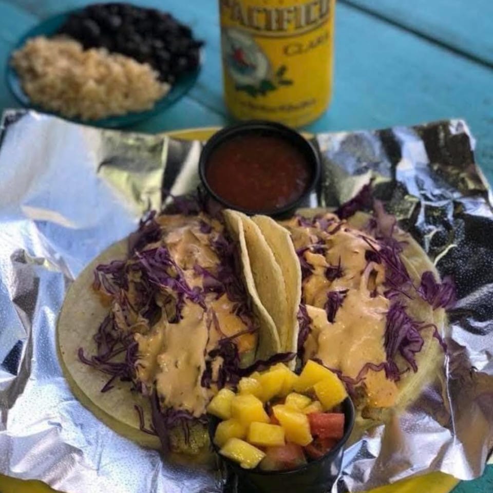 Two tacos with purple cabbage, creamy sauce, fruit salsa, and a beer on a blue table.