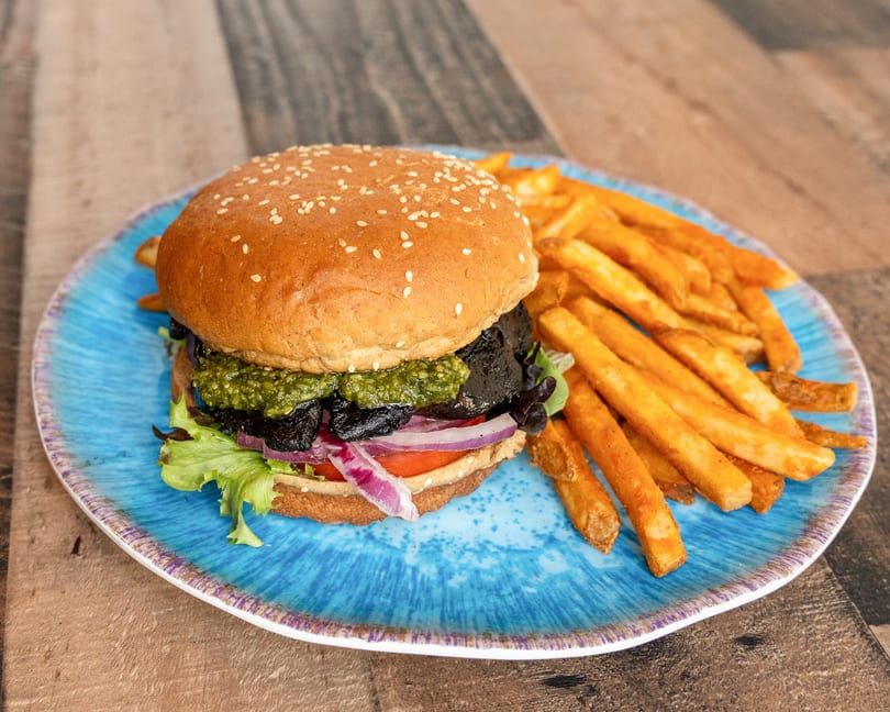 A veggie burger with pesto, mushrooms, red onion, lettuce, and tomato, served with fries on a blue plate.