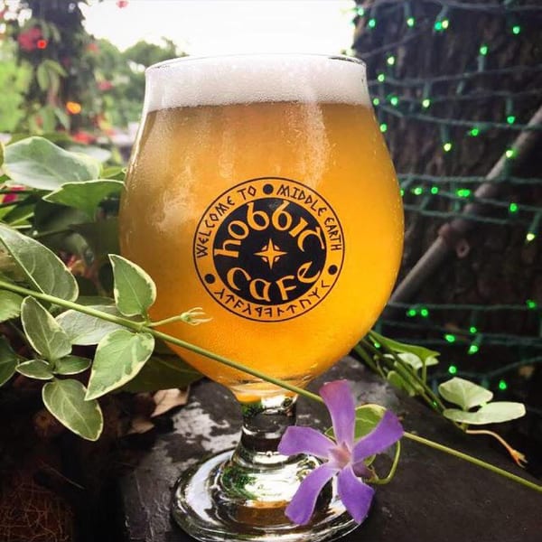 Hobbit Cafe branded beer glass filled with golden ale, surrounded by green foliage and a purple flower.