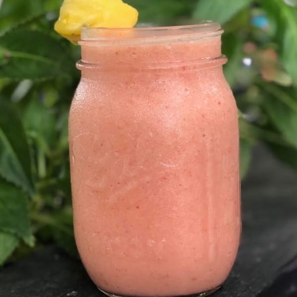 Pink smoothie in a mason jar with a pineapple garnish, against a leafy green background.