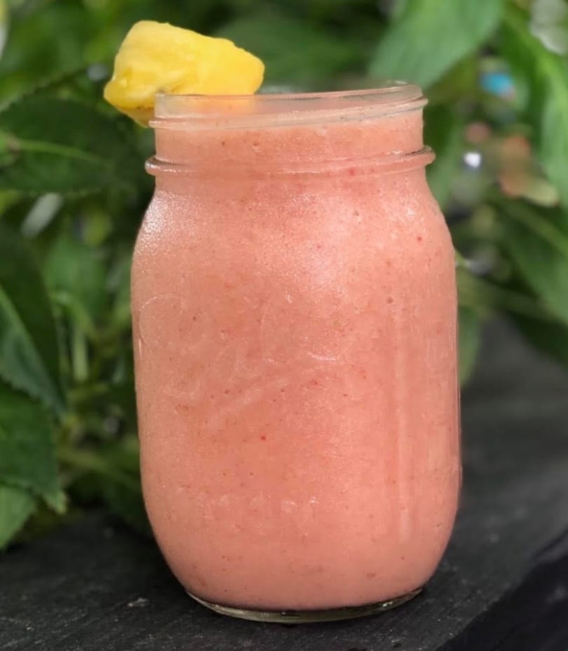 Pink smoothie in a mason jar with a pineapple garnish, against a leafy green background.