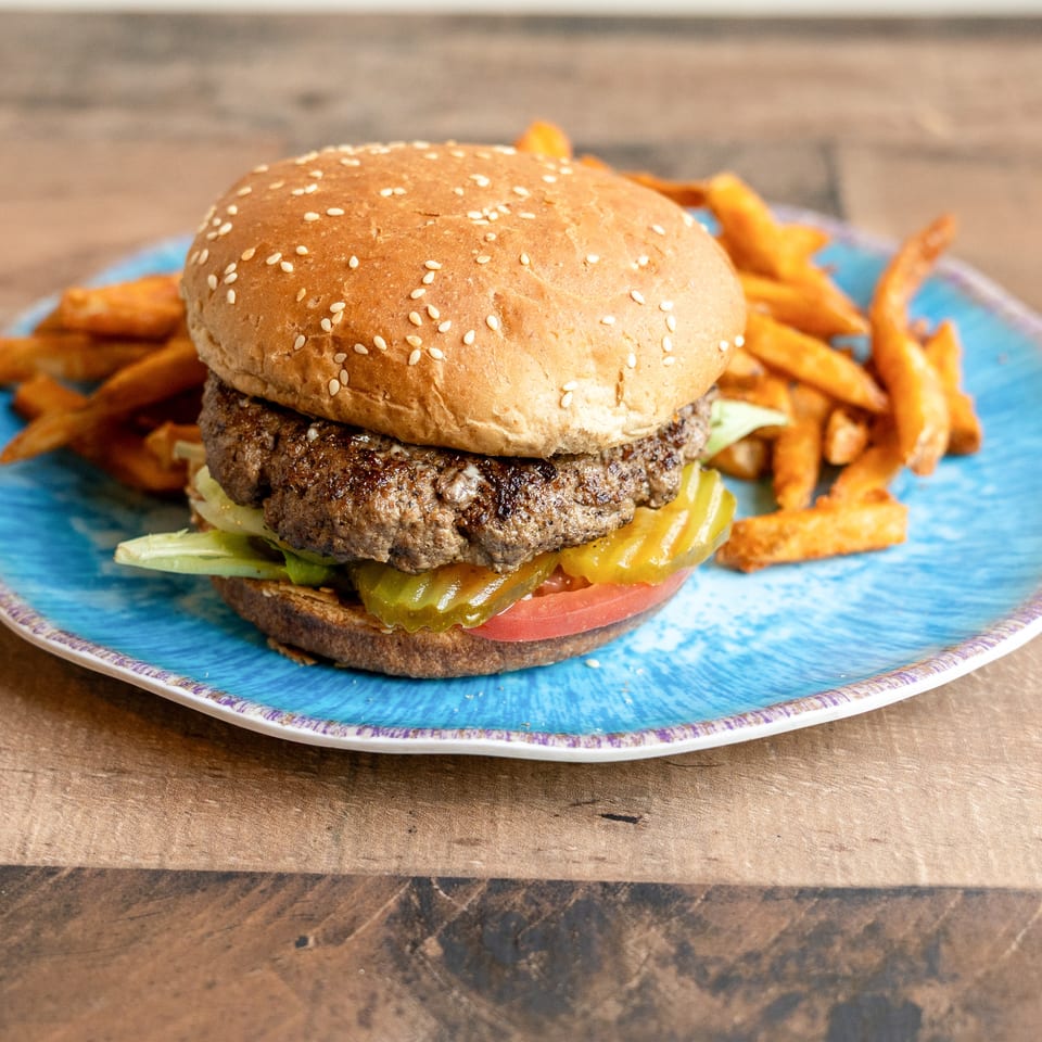 A juicy hamburger with lettuce, tomato, and pickles, served with sweet potato fries on a blue plate.