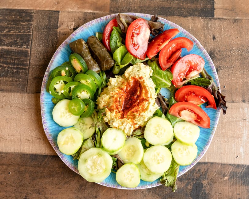 A vibrant platter with hummus, dolmas, sliced tomatoes, cucumbers, and jalapeños on mixed greens.
