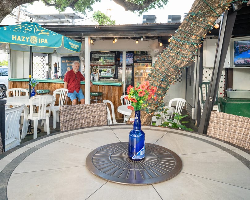 An outdoor bar with a man in a red shirt, white tables, and a tree wrapped in lights.