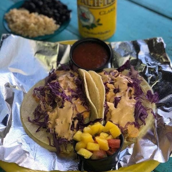 Two tacos with purple cabbage and sauce, salsa, fruit, rice, beans, and a beer can.