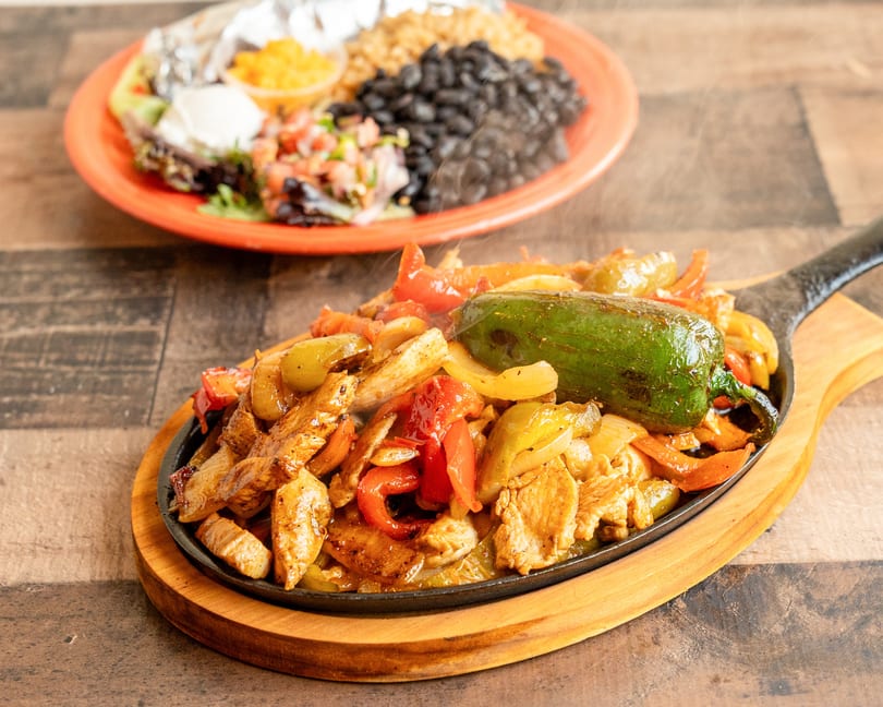 Sizzling chicken fajitas with peppers and onions on a cast iron skillet, with a side plate of rice and beans.