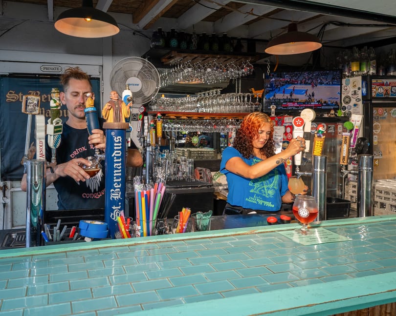 Two bartenders pour drinks behind a teal-tiled bar, with beer taps and glassware visible.